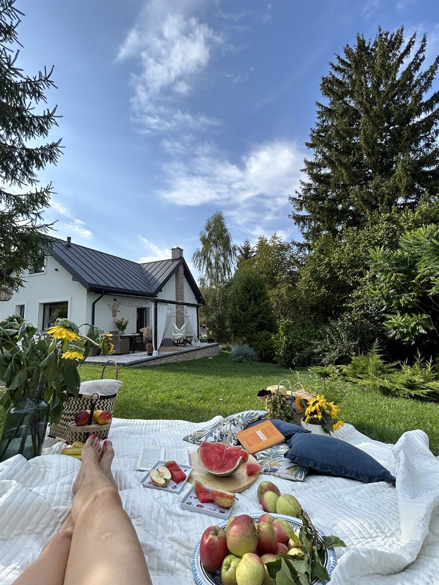 piknikowy koc na trawie z owocami (arbuz, jabłka), poduszkami i nogami w stronę białego domu i drzew, słoneczny dzień