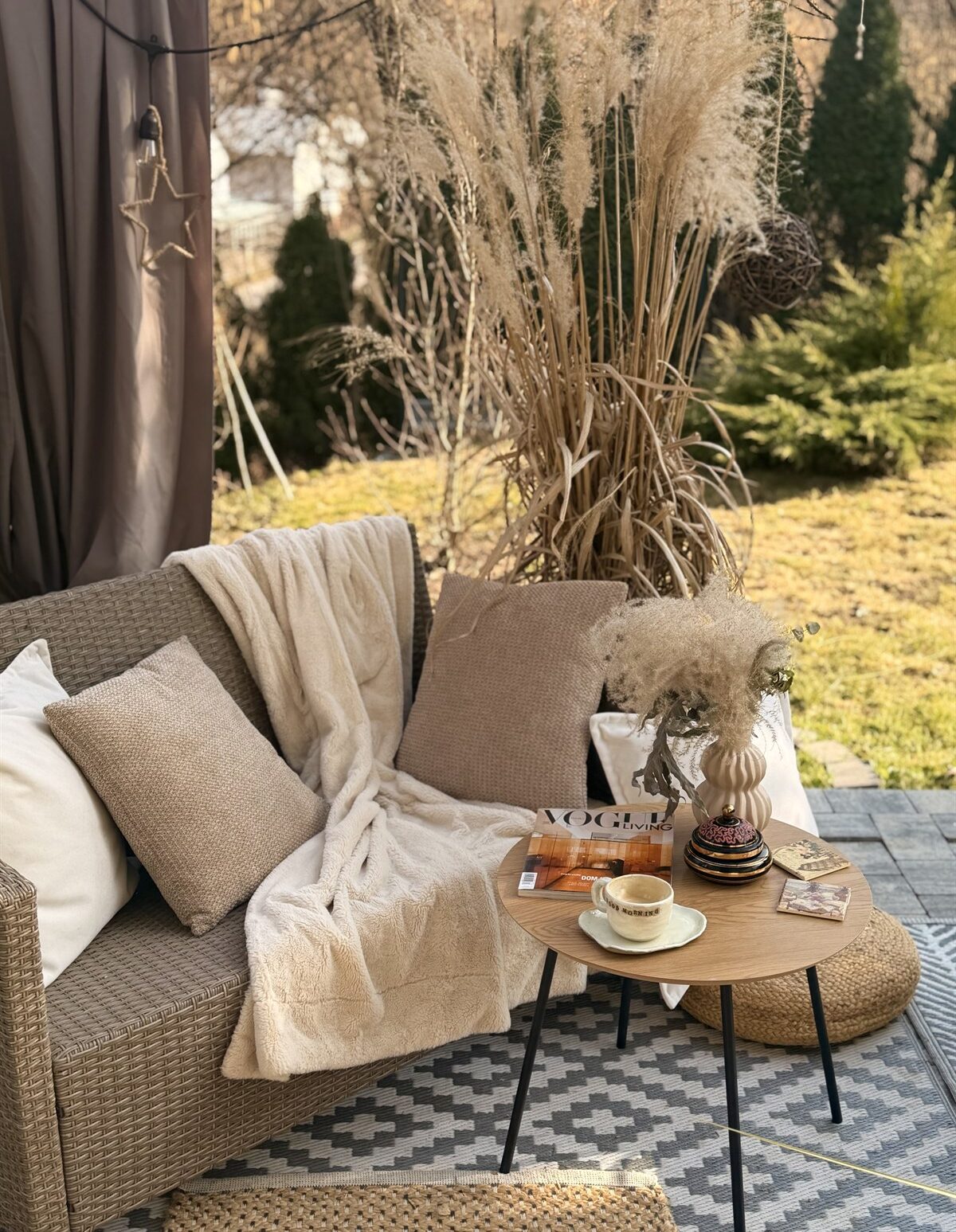 Przytulny kącik na tarasie z rattanową sofą, beżowymi poduszkami, kocem i stolikiem z filiżanką i magazynem VOGUE
