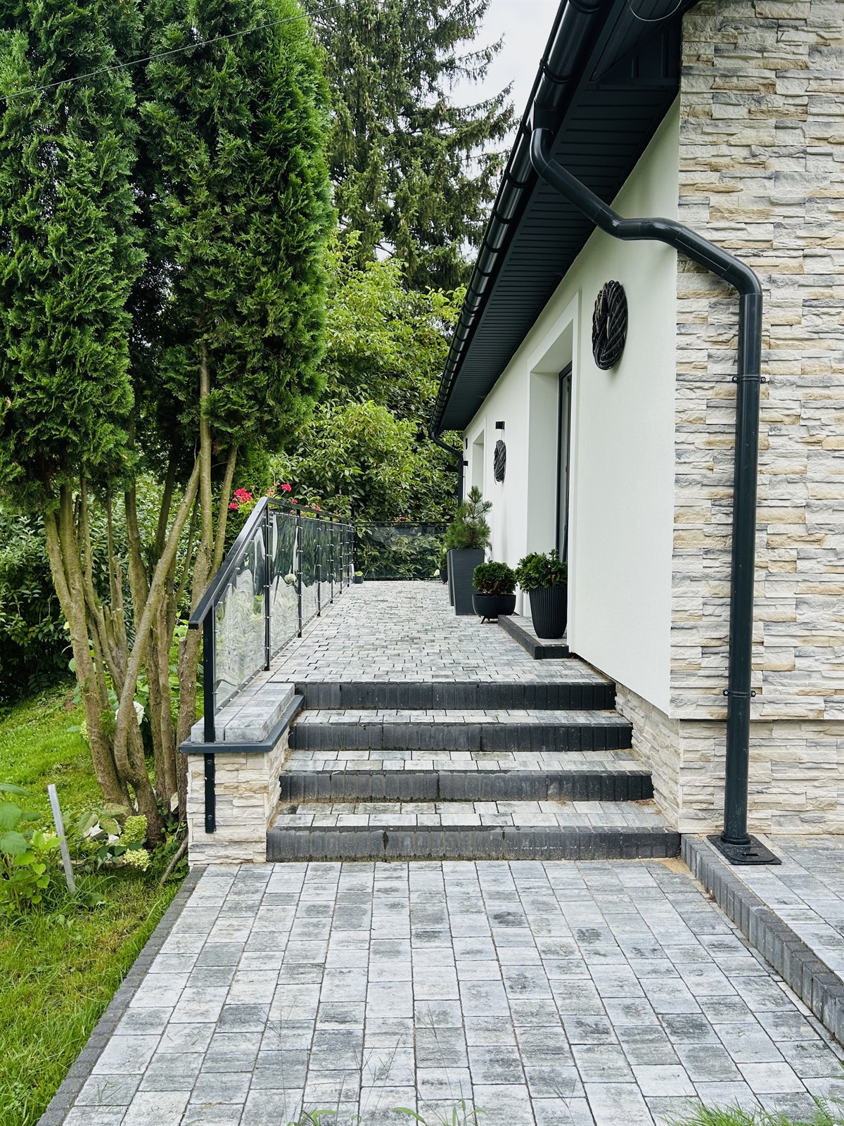 Wejście do nowoczesnego domu z szarymi kamiennymi stopniami, szklaną balustradą i donicami przy ścianie