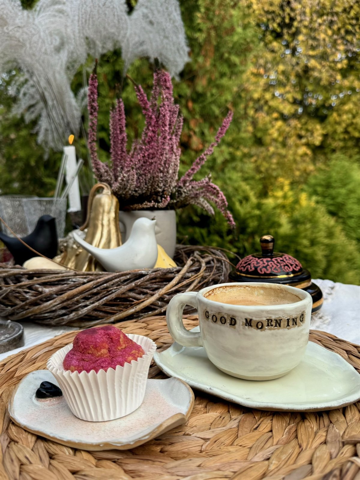 Filiżanka kawy z napisem GOOD MORNING obok różowego muffinka i wrzosu w koszu w ogrodowej scenerii